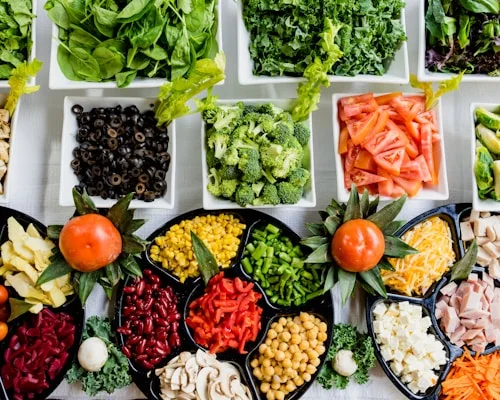 An overhead view of a salad bar: bowls of spinach, kale, broccoli, tomatoes, peppers, sweetcorn, beans, chickpeas and mushrooms